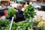 A person in a headscarf holds up two bunches of leafy greens in a vegetable market